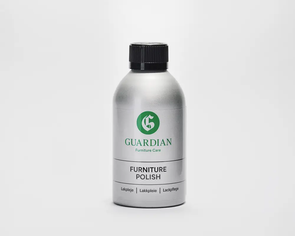 En sølvfarvet flaske med sort skruelåg, der har et grønt 'G'-logo og mærket 'Guardian' på forsiden. Etiketten viser også teksten 'Furniture Care' og 'Furniture Polish' med sort skrift. Flasken har et slankt, moderne design og ser ud til at være lavet af metal eller metallisk plast. Nederst på etiketten står produktinformation på flere sprog.