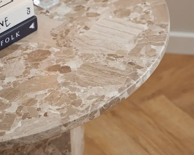 Nærbillede af en rund bordplade med en poleret overflade af natursten, der har et miks af beige, lysebrune og cremefarvede nuancer i et uregelmæssigt mønster. Kanten er glat og bordpladen har en blank finish. Et par bøger er delvist synlige i venstre side af billedet.