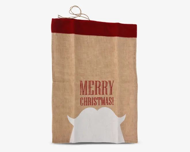 Rektangulær stofpose med naturlig jutestruktur og en bred rød kant øverst. Forsiden har teksten 'MERRY CHRISTMAS!' trykt med store røde bogstaver. Under teksten er der en hvid silhuet af et julemandskæg og overskæg. Posen har en lille løkke øverst til ophængning.