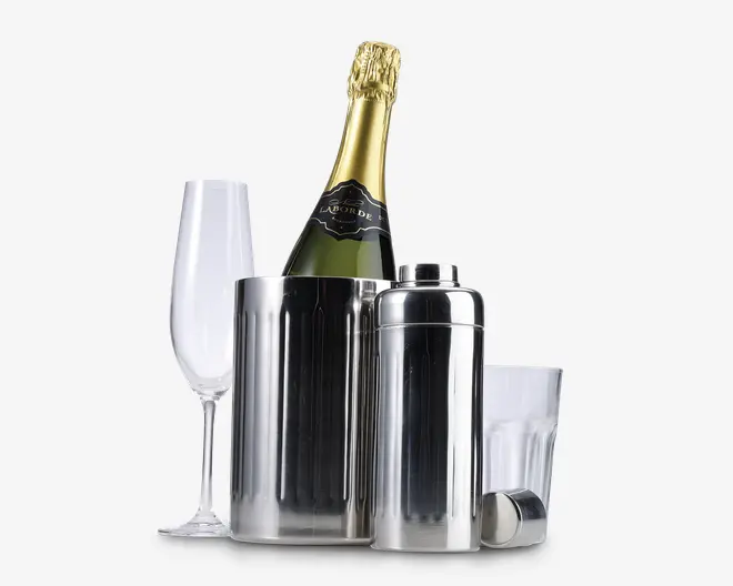 Billedet viser et sæt barudstyr i poleret rustfrit stål, herunder en høj isspand med lodrette riller, en cocktailshaker med tilhørende låg og et klart glas tumbler. Der er også et champagneglas af klart glas. Is-spanden indeholder en flaske champagne med gyldent folie på toppen og sort etiket. Udseendet er moderne og elegant, velegnet til servering af drikkevarer til fest eller sammenkomst.