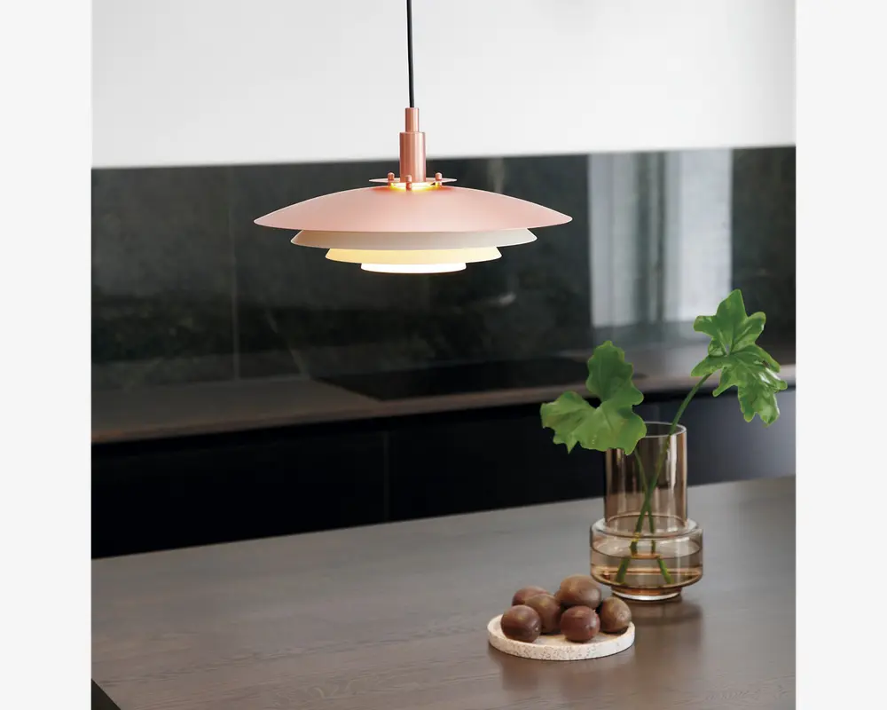 En moderne pendellampe med en lagdelt, skiveformet skærm i en blød rosa farve. Lampen hænger i en sort ledning og udsender et varmt, diffust lys nedad. Designet er minimalistisk og nutidigt, velegnet til køkkener eller spisestuer. Lampens overflade virker mat, og den lagdelte konstruktion reducerer blænding og giver jævn belysning.