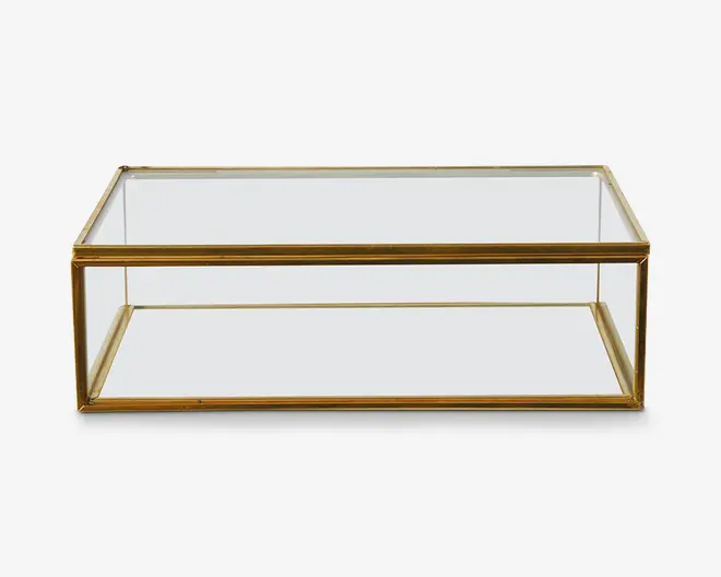 Rektangulært bord med klar glasplade og en nederste hylde, understøttet af en slank metalramme i guldfarve. Designet er minimalistisk og moderne med rene linjer og en åben struktur, der giver både udstillings- og opbevaringsplads.