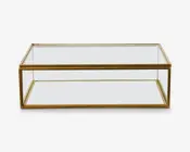 Rektangulært bord med klar glasplade og en nederste hylde, understøttet af en slank metalramme i guldfarve. Designet er minimalistisk og moderne med rene linjer og en åben struktur, der giver både udstillings- og opbevaringsplads.
