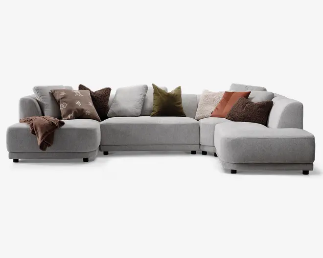 En stor, moderne hjørnesofa betrukket med lysegråt stof. Sofaen har brede, dybe sæder og lave, afrundede armlæn. Den er arrangeret i en L-form med en chaiselong på højre side. Sofaen er pyntet med forskellige pyntepuder i jordfarver, herunder brun, olivengrøn og rustorange, samt et brunt tæppe, der er lagt over venstre armlæn. Det samlede udtryk er hyggeligt og indbydende, velegnet til en moderne stue.