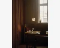 En hyggelig restaurantbås med en brun læderbænk og en træstol, placeret op ad en væg med varme brune gardiner. Bordet er dækket til to personer med et vinglas, et vandglas og pænt arrangeret bestik på hvide servietter. Over bordet hænger en moderne rund pendellampe, der giver et blødt lys. Et vindue i baggrunden lukker dagslys ind, og der står flasker i vindueskarmen, hvilket bidrager til den indbydende stemning.