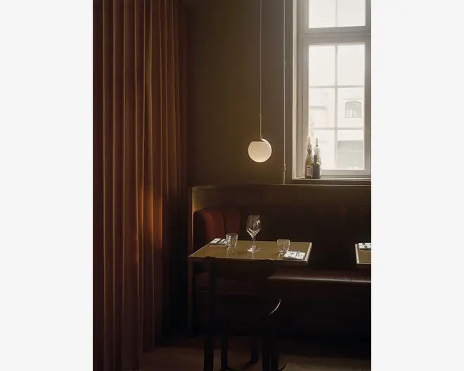 En hyggelig restaurantbås med en brun læderbænk og en træstol, placeret op ad en væg med varme brune gardiner. Bordet er dækket til to personer med et vinglas, et vandglas og pænt arrangeret bestik på hvide servietter. Over bordet hænger en moderne rund pendellampe, der giver et blødt lys. Et vindue i baggrunden lukker dagslys ind, og der står flasker i vindueskarmen, hvilket bidrager til den indbydende stemning.