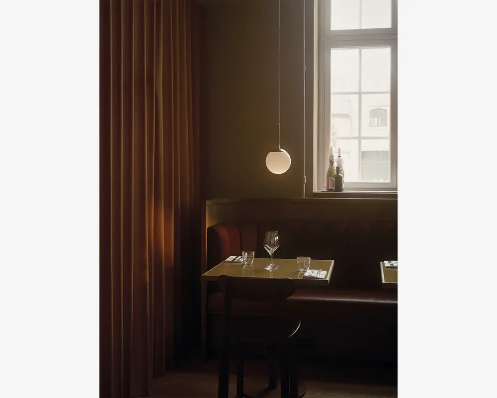 En hyggelig restaurantbås med en brun læderbænk og en træstol, placeret op ad en væg med varme brune gardiner. Bordet er dækket til to personer med et vinglas, et vandglas og pænt arrangeret bestik på hvide servietter. Over bordet hænger en moderne rund pendellampe, der giver et blødt lys. Et vindue i baggrunden lukker dagslys ind, og der står flasker i vindueskarmen, hvilket bidrager til den indbydende stemning.