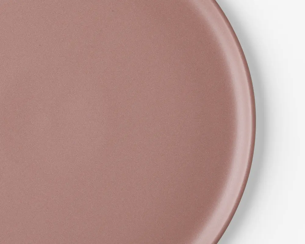 Et nærbillede af en rund tallerken med en glat, mat overflade i en afdæmpet rosa farve. Tallerkenen har en let forhøjet kant og en ensartet overflade, hvilket giver den et moderne og minimalistisk udtryk.