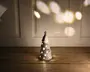 En lille keramisk pyntegenstand formet som et juletræ, malet hvid med en blank overflade. Træet har udskårne huller og stjerneformer, hvorigennem et varmt hvidt lys skinner indefra og skaber en blød, glødende effekt. Øverst på træet sidder en gylden stjerne, og der er malet fine grene og bær på overfladen. Pyntegenstanden står på en træoverflade, og lyset danner dekorative mønstre på væggen bagved.