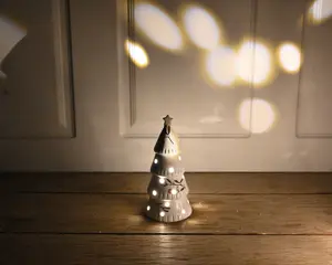 En lille keramisk pyntegenstand formet som et juletræ, malet hvid med en blank overflade. Træet har udskårne huller og stjerneformer, hvorigennem et varmt hvidt lys skinner indefra og skaber en blød, glødende effekt. Øverst på træet sidder en gylden stjerne, og der er malet fine grene og bær på overfladen. Pyntegenstanden står på en træoverflade, og lyset danner dekorative mønstre på væggen bagved.