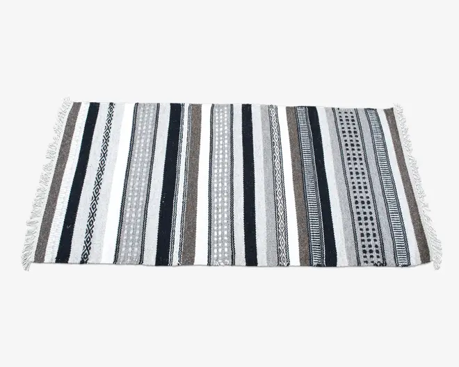 Rektangulært tekstil med skiftende lodrette striber i sorte, hvide, grå og brune nuancer. Striberne har forskellige mønstre, herunder ensfarvede, geometriske former og prikdesign. Begge korte sider er afsluttet med hvide frynser.