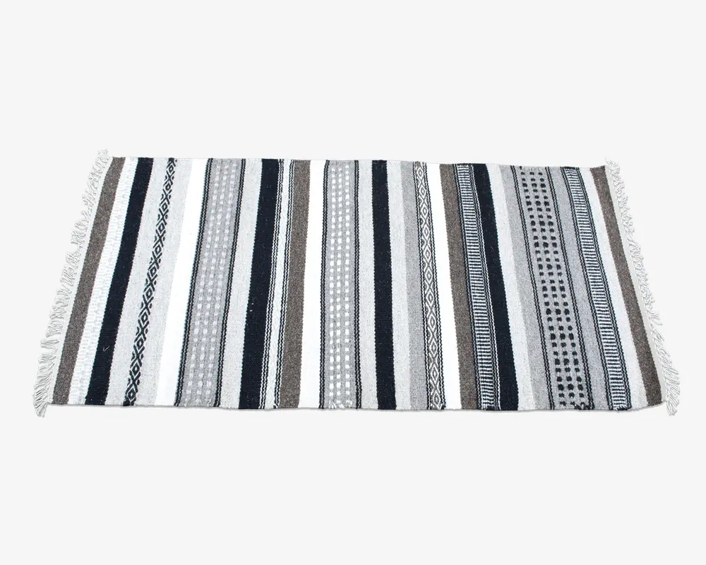 Rektangulært tekstil med skiftende lodrette striber i sorte, hvide, grå og brune nuancer. Striberne har forskellige mønstre, herunder ensfarvede, geometriske former og prikdesign. Begge korte sider er afsluttet med hvide frynser.