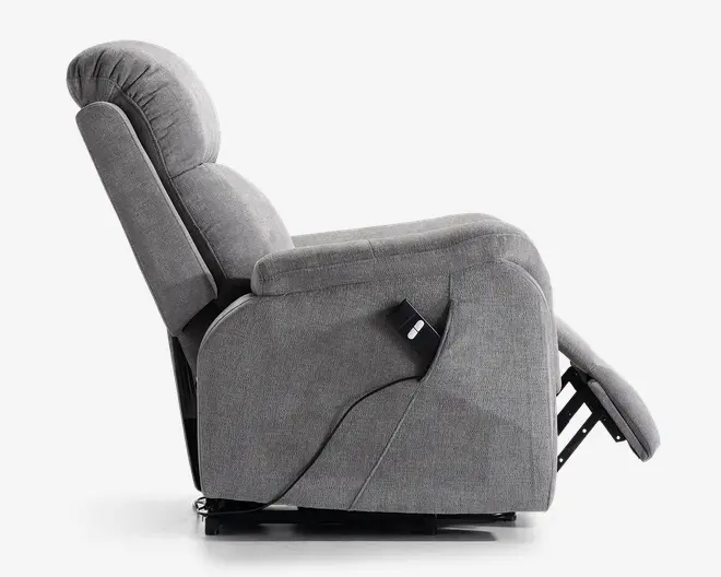 Sidebillede af en grå polstret lænestol i fuldt tilbagelænet position. Stolen har en kraftigt polstret ryg, sæde og armlæn for ekstra komfort. En ledningsforbundet fjernbetjening er fastgjort på siden, hvilket indikerer elektrisk tilbagelæningsfunktion. Fodstøtten er slået ud, og designet er moderne og blødt.
