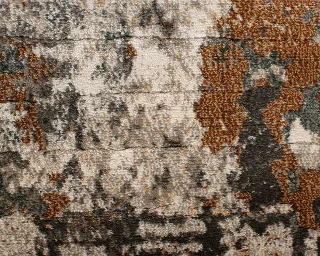 Nærbillede af et tekstiler med en abstrakt, slidt mønster. Overfladen har en blanding af jordfarver som brun, beige, grå og strejf af blågrøn. Designet fremstår ujævnt og plettet med områder af både høj og lav luv, hvilket giver en taktil og tredimensionel fornemmelse. Farverne er blandet på en uregelmæssig, organisk måde, hvilket skaber et moderne og kunstnerisk udtryk.