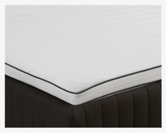 Nærbillede af en madras med et hvidt, quiltet og let polstret toplag med diskrete horisontale syninger og mørkegrå kantbånd. Madrassens bund er mørk med lodrette syninger, hvilket giver en kontrast til det lyse, rene toplag.