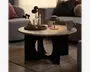 Rund bordplade i stenlook med glat lys marmoreret finish og en markant sort skulpturel base med en stor oval udskæring. Stylet i en hyggelig stue med stearinlys, krus og en lille bordlampe ovenpå.