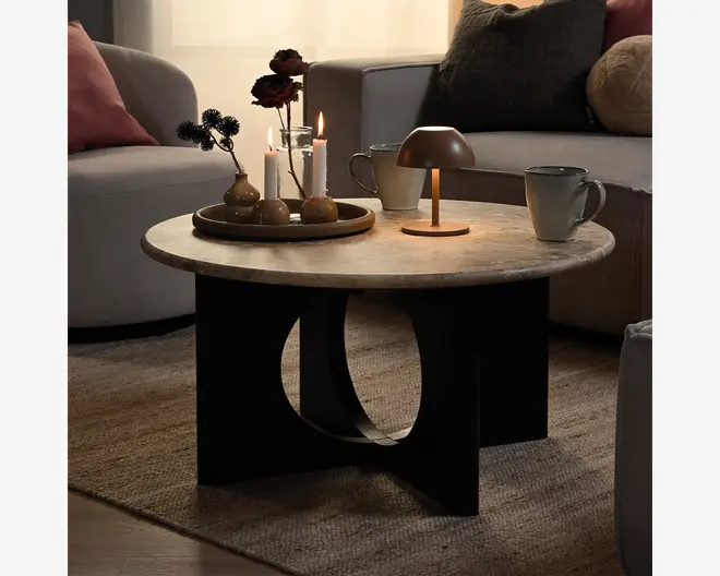 Rund bordplade i stenlook med glat lys marmoreret finish og en markant sort skulpturel base med en stor oval udskæring. Stylet i en hyggelig stue med stearinlys, krus og en lille bordlampe ovenpå.