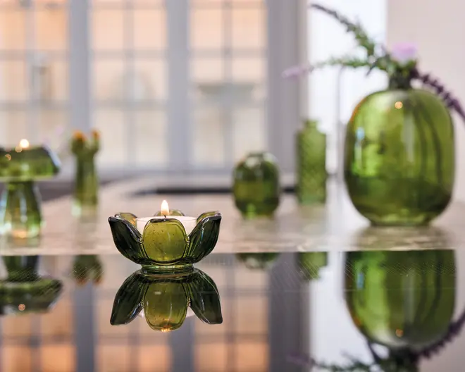 En grøn fyrfadsstage i glas formet som en blomst med en tændt lys indeni står på en reflekterende overflade. Glasset er gennemsigtigt med kronbladslignende kanter. I baggrunden ses forskellige grønne glasvaser og beholdere i forskellige former og størrelser, nogle med dekorative blomster. Stemningen er elegant og moderne med blødt, stemningsfuldt lys.