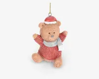 En sød bamsefigur med tekstureret pels, iført en rød strikket sweater, en rød nissehue med hvid kant og et lysegråt tørklæde. Bamsen sidder med armene løftet og har et venligt udtryk. Der er en gylden ophængssnor fastgjort i toppen til nem ophængning.