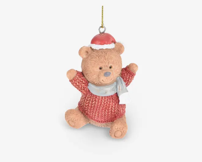 En sød bamsefigur med tekstureret pels, iført en rød strikket sweater, en rød nissehue med hvid kant og et lysegråt tørklæde. Bamsen sidder med armene løftet og har et venligt udtryk. Der er en gylden ophængssnor fastgjort i toppen til nem ophængning.