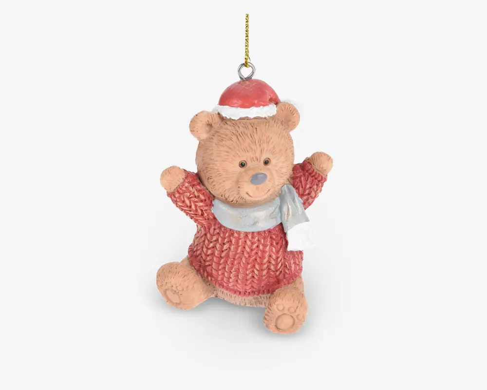 En sød bamsefigur med tekstureret pels, iført en rød strikket sweater, en rød nissehue med hvid kant og et lysegråt tørklæde. Bamsen sidder med armene løftet og har et venligt udtryk. Der er en gylden ophængssnor fastgjort i toppen til nem ophængning.
