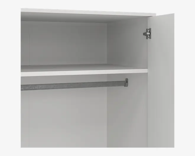 Et nærbillede af indersiden af et skab med hvid finish. Skabet har en enkelt hylde placeret i den øverste halvdel, hvilket giver opbevaringsplads ovenover. Under hylden er der en solid metalstang til ophængning af tøj. Skabslågen er åben, og der ses et metalhængsel på højre side. Designet er enkelt og moderne med rene linjer og en glat overflade.