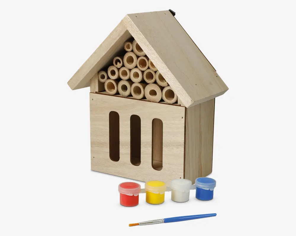 En husformet struktur af træ med skråt tag, der har flere hule bambusrør arrangeret under taget og tre lodrette åbninger på frontpanelet. Træet er ubehandlet, hvilket gør det muligt at dekorere. Foran strukturen ligger fire små malerbøtter i rød, gul, hvid og blå samt en pensel, hvilket viser, at produktet kan males og personliggøres.