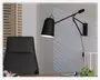 Mat sort væglampe med justerbar arm og vinklet skærm, designet til retningsbestemt belysning. Lampen har et moderne, minimalistisk udtryk og er tilsluttet en stikkontakt med en synlig ledning. Den er placeret over et skrivebord og giver målrettet lys til arbejdsopgaver.