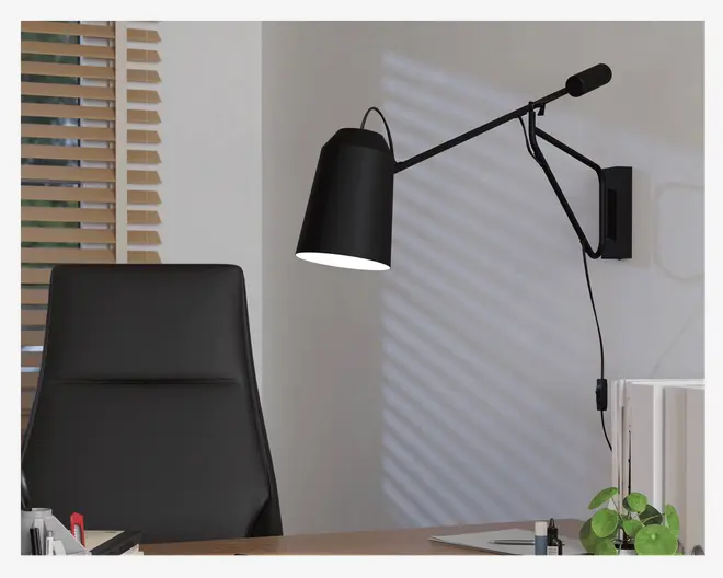 Mat sort væglampe med justerbar arm og vinklet skærm, designet til retningsbestemt belysning. Lampen har et moderne, minimalistisk udtryk og er tilsluttet en stikkontakt med en synlig ledning. Den er placeret over et skrivebord og giver målrettet lys til arbejdsopgaver.