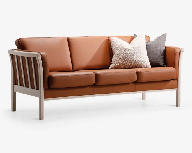 En tresæders sofa med lysebrune læderhynder og en ramme af lyst træ. Sofaen har lameller i træ på siderne og bagpå, og der medfølger to pyntepuder: én stor, fluffy pude i cremefarvet og én mindre, tekstureret pude i mørkegrå. Sæde- og ryghynderne er tykke og bløde, hvilket giver god siddekomfort.