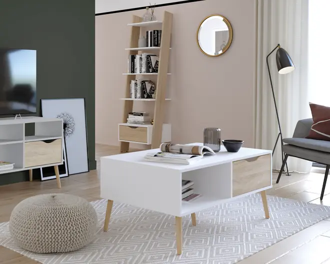 Rektangulært sofabord i moderne skandinavisk design med hvid bordplade og åbne opbevaringsrum på begge sider. Bordet står på fire skrå ben i lyst træ. På bordpladen står et glas, en skål og et åbent magasin. Bordet er placeret på et lyst tæppe med geometrisk mønster i en lys stue.