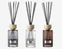 Sæt med tre elegante duftpinde i glasflasker med lodrette riller og en metallabel på forsiden. Flaskerne har tre forskellige farver: røggrå, klar og varm brun. Hver diffuser har et låg af lyst træ og indeholder flere sorte pinde til at sprede duften. Designet er moderne og stilfuldt, velegnet til bolig- eller kontorindretning.