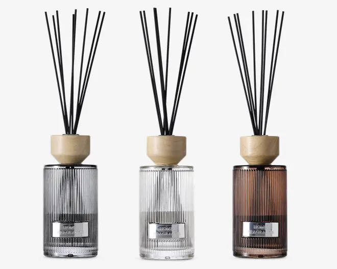 Sæt med tre elegante duftpinde i glasflasker med lodrette riller og en metallabel på forsiden. Flaskerne har tre forskellige farver: røggrå, klar og varm brun. Hver diffuser har et låg af lyst træ og indeholder flere sorte pinde til at sprede duften. Designet er moderne og stilfuldt, velegnet til bolig- eller kontorindretning.