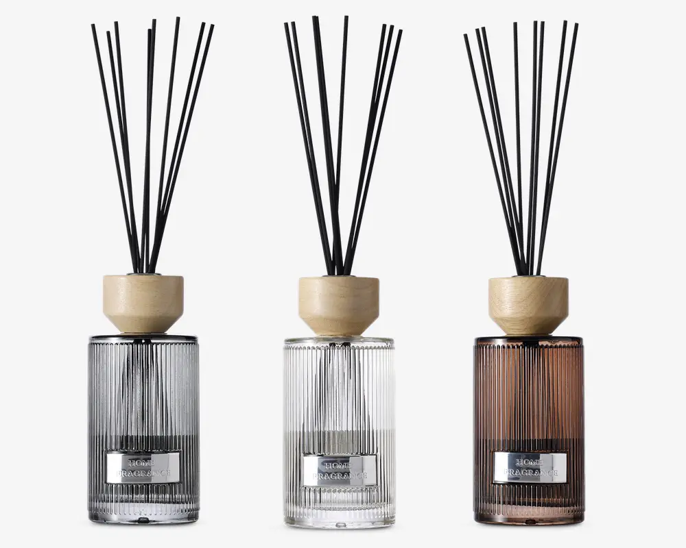 Sæt med tre elegante duftpinde i glasflasker med lodrette riller og en metallabel på forsiden. Flaskerne har tre forskellige farver: røggrå, klar og varm brun. Hver diffuser har et låg af lyst træ og indeholder flere sorte pinde til at sprede duften. Designet er moderne og stilfuldt, velegnet til bolig- eller kontorindretning.