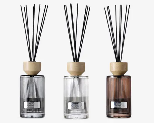 Sæt med tre elegante duftpinde i glasflasker med lodrette riller og en metallabel på forsiden. Flaskerne har tre forskellige farver: røggrå, klar og varm brun. Hver diffuser har et låg af lyst træ og indeholder flere sorte pinde til at sprede duften. Designet er moderne og stilfuldt, velegnet til bolig- eller kontorindretning.