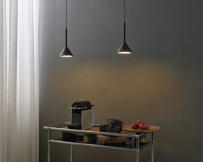 Et moderne køkkenarrangement med en kompakt espressomaskine i stilrent metalfinish, en sort kaffekop med underkop og en matchende sort mælkekande. Disse genstande er placeret på en træbakke oven på en minimalistisk metalvogn. Baggrunden består af en glat, mørk væg, der oplyses af to sorte, kegleformede pendellamper, som hænger fra loftet og skaber en hyggelig stemning. Et par røde grydelapper ligger også på bakken og tilføjer et farverigt element til scenen.