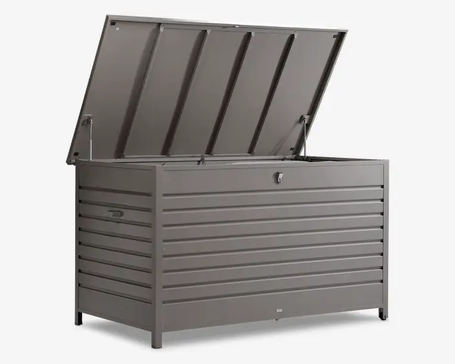 Stor, rektangulær udendørs opbevaringsboks lavet af metal med mat grå finish. Boksen har horisontale lameller på siderne og fronten for ekstra styrke og et stilrent udseende. Låget er hængslet og holdes åbent med gasdæmpere for nem adgang, og der er en lås på fronten for sikkerhed. Håndtag på siderne gør det lettere at flytte boksen.