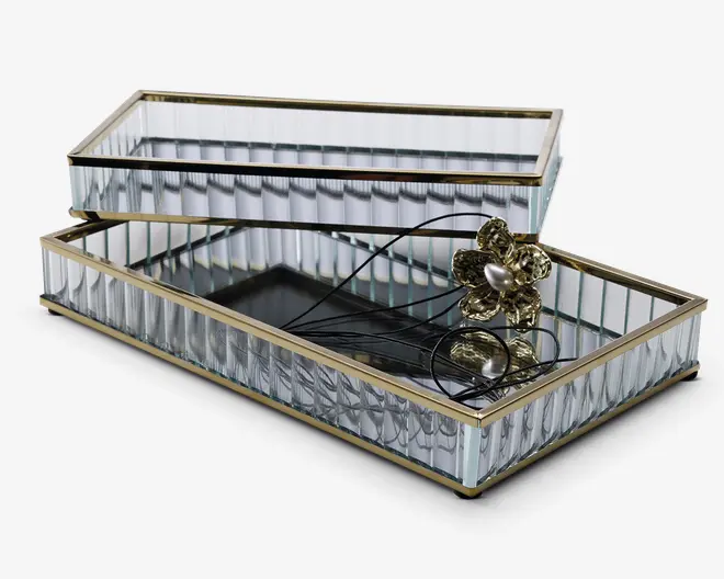Rektangulær glasæske med hængslet låg, der har lodrette riflede glassider og guldfarvet metalramme langs kanterne. Æsken har en spejlbund og indeholder et guldfarvet blomsterformet vedhæng med en perle i midten på en sort snor.