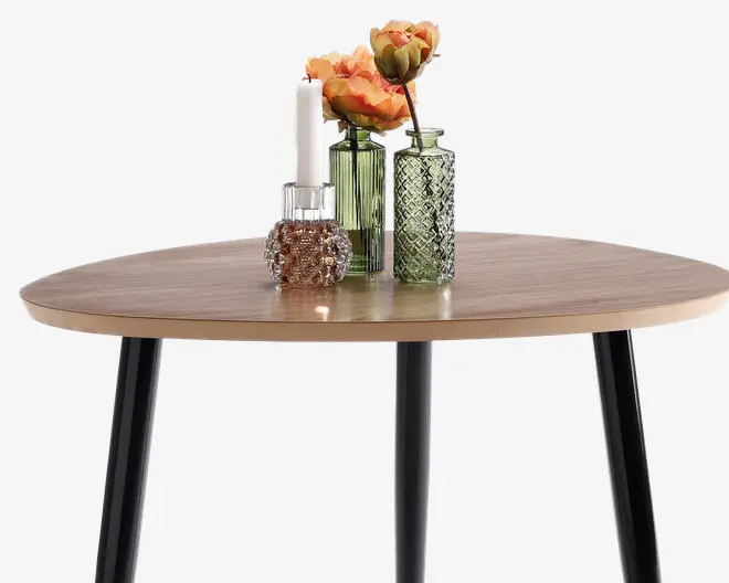 Rund bordplade med lys træfinish og sorte skrå ben. Bordet er pyntet med en grøn rillet glasvase med orange kunstige blomster, en grøn mønstret glasvase og en lille klar glaslysestage med et hvidt stearinlys. Opsætningen giver et moderne og elegant udtryk.