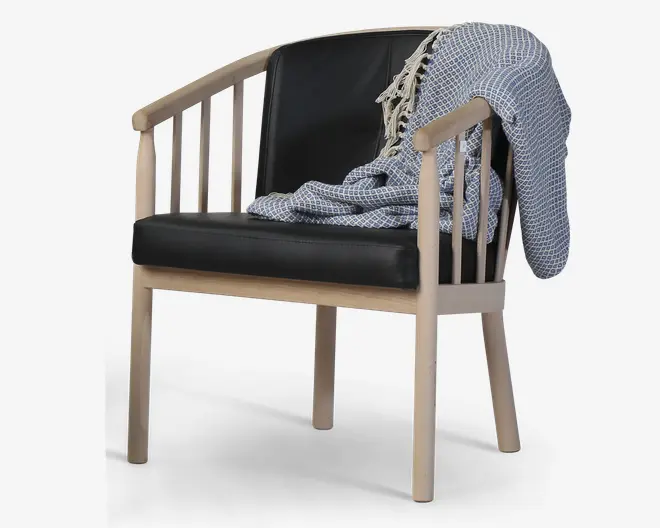 En moderne lænestol med en lys træramme og lodrette lameller på siderne og ryggen. Sædet og ryglænet er polstret med glat sort læder. Over den ene armlæn ligger et blåt og hvidt mønstret tæppe med frynser, som giver stolen et hyggeligt udtryk.