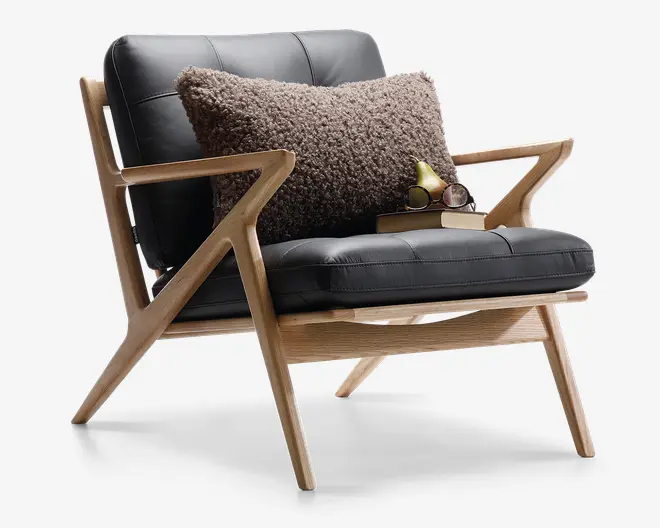 Stol med stel i lyst træ og et vinklet, retro-inspireret design. Sæde og ryglæn er polstret med sort læder eller læderlignende materiale med synlige syninger. En stor, tekstureret brun pude ligger på ryglænet og giver ekstra komfort og stil. På sædet ligger to stablede bøger, et par runde briller og en dekorativ pære, hvilket skaber en hyggelig læsekrog.