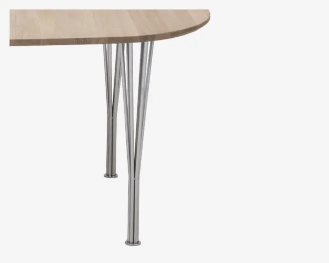 Billedet viser et nærbillede af et bordhjørne med en bordplade i lyst træ og afrundet kant. Bordet understøttes af tre slanke, skråtstillede metalben med en blank kromfinish, hvilket giver et moderne og minimalistisk udtryk.