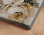 Nærbillede af et lærredsprint med et tekstureret, håndmalet blomsterdesign i nuancer af creme, beige og guld. Penselstrøgene er tydelige og giver kunstværket et tredimensionelt, kunstnerisk udtryk. Lærredet er stramt spændt omkring en træramme med skarpe, rene kanter. Baggrunden viser et trægulv med en lys, naturlig finish.