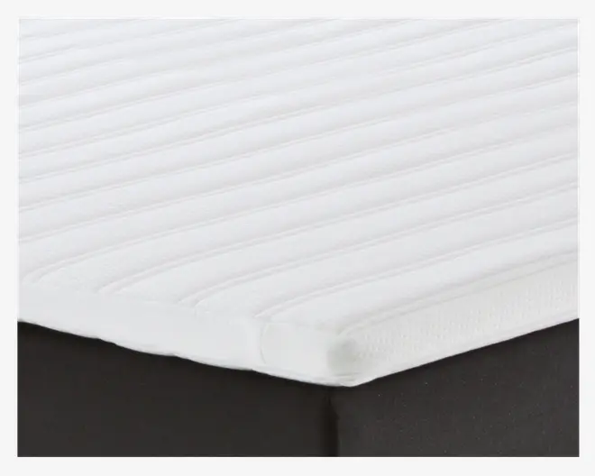 Nærbillede af en madras-topper med hvid, tekstureret overflade og jævnt fordelte vandrette riller. Topperen er tynd og ligger tæt oven på madrassen, hvilket giver et ekstra komfortlag. Kanterne er pænt afsluttede, og materialet ser blødt og åndbart ud.