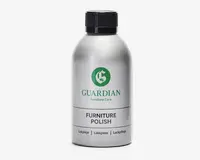 En sølvfarvet flaske med sort skruelåg, der har et grønt 'G'-logo og mærket 'Guardian Furniture Care' på etiketten. Etiketten viser også produktnavnet 'Furniture Polish' med fed sort skrift samt oversættelser på dansk, norsk og tysk. Flasken har et stilrent, moderne design og ser ud til at være lavet af metal eller metallisk plast.