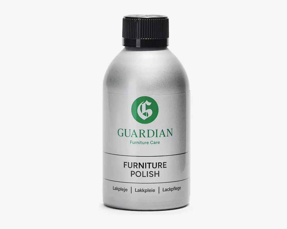 En sølvfarvet flaske med sort skruelåg, der har et grønt 'G'-logo og mærket 'Guardian Furniture Care' på etiketten. Etiketten viser også produktnavnet 'Furniture Polish' med fed sort skrift samt oversættelser på dansk, norsk og tysk. Flasken har et stilrent, moderne design og ser ud til at være lavet af metal eller metallisk plast.