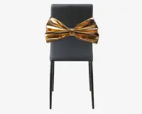 En moderne stol med en elegant, mat sort finish og slanke, lige ben. Det mest iøjnefaldende træk er en stor, metallisk guldfarvet sløjfe, der er fastgjort til ryglænet og giver stolen et markant og luksuriøst udseende. Sløjfen er blank og formet, hvilket tilføjer et dekorativt og opsigtsvækkende element til det ellers minimalistiske design.
