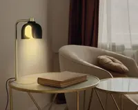 En moderne bordlampe med en guldfarvet metalbase og en buet arm, der holder en sort, kuppelformet lampeskærm. Lampen udsender et varmt, blødt gult lys. Den står på et rundt sidebord med glasplade og guldfarvet kant, ved siden af en lukket bog med hårdt omslag. I baggrunden ses en beige polstret lænestol med en brun pude samt et vindue med hvide gardiner og mørkebrune draperier.