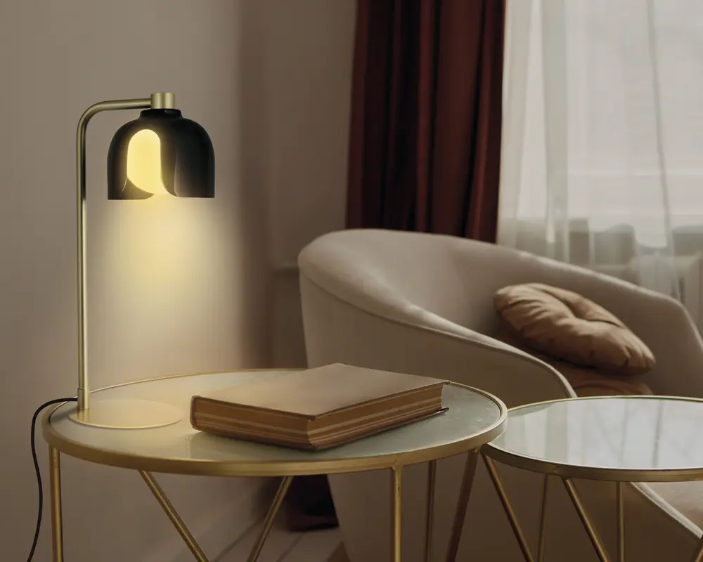 En moderne bordlampe med en guldfarvet metalbase og en buet arm, der holder en sort, kuppelformet lampeskærm. Lampen udsender et varmt, blødt gult lys. Den står på et rundt sidebord med glasplade og guldfarvet kant, ved siden af en lukket bog med hårdt omslag. I baggrunden ses en beige polstret lænestol med en brun pude samt et vindue med hvide gardiner og mørkebrune draperier.