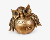 En dekorativ uglefigur lavet af metal med en gylden finish. Uglen har en rund, glat krop med indgraverede fjerdetaljer og store, sorte, blanke øjne. Vingerne er teksturerede og strakt let ud til siderne, og der er små skulpturerede bær nær basen. Det samlede design er finurligt og elegant, velegnet til boligindretning.